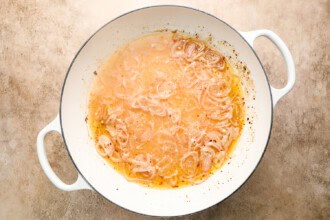 Image for recipe instruction: Sliced onions cooking in a white enameled pot with liquid, creating softened onions and a golden broth reminiscent of classic Moules Frites preparations.