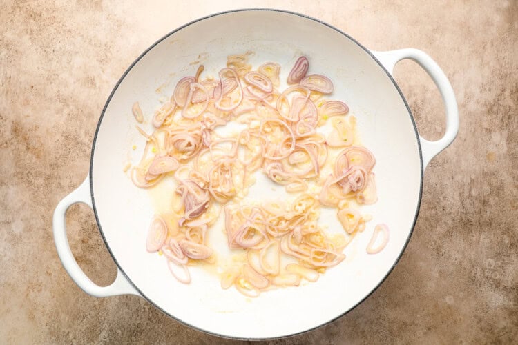 Image for recipe instruction: A white pan on a beige surface contains thinly sliced shallots sautéing in melted butter, evoking the flavors of Belgian cuisine and traditional Moules Frites.