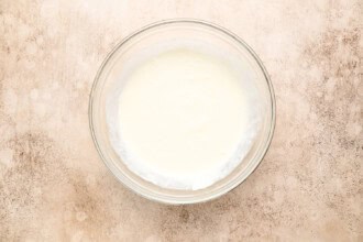 Image for recipe instruction: A clear glass bowl filled with plain white yogurt sits on a beige textured surface, adding a simple contrast to the rich flavors of Moules Frites.