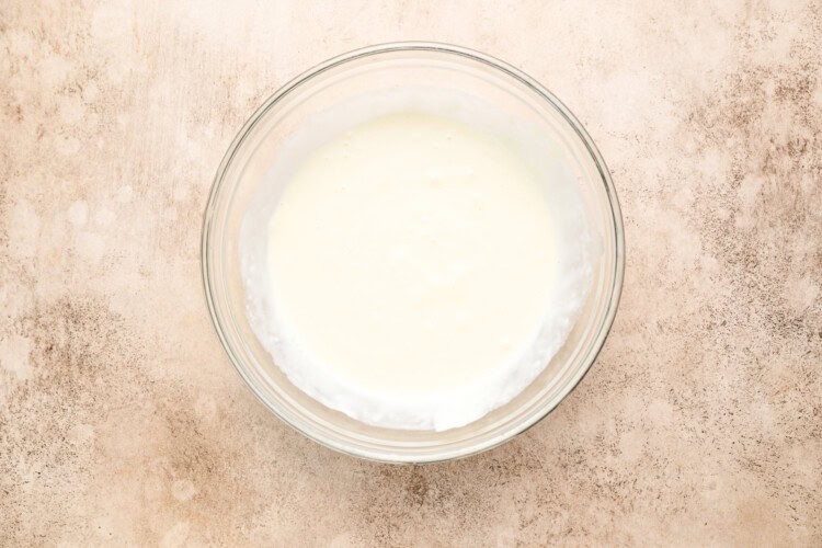 Image for recipe instruction: A clear glass bowl filled with plain white yogurt sits on a beige textured surface, adding a simple contrast to the rich flavors of Moules Frites.