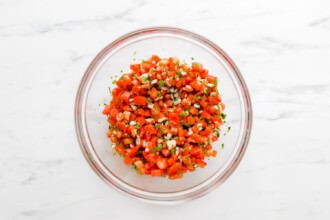 Image for recipe instruction: A clear glass bowl of pico de gallo, brimming with chopped tomatoes, onions, cilantro, and jalapeños, sits on a white marble surface.
