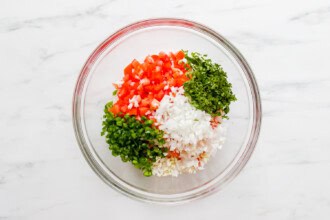 Image for recipe instruction: A glass bowl with fresh Pico de Gallo, featuring chopped tomatoes, onions, cilantro, jalapeños, and garlic on a white marble surface.