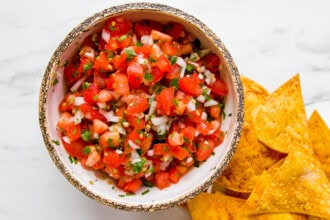 Image for recipe instruction: A bowl of fresh Pico de Gallo salsa with chopped tomatoes, onions, and herbs sits beside a pile of crispy tortilla chips on a white surface.