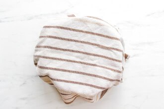 Image for recipe instruction: A bowl of Pico de Gallo covered with a white kitchen towel featuring brown stripes, placed on a white marble surface.