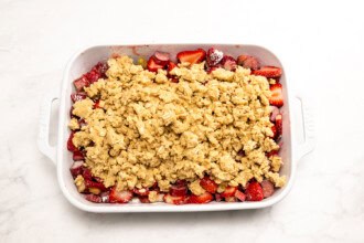 Image for recipe instruction: A white baking dish filled with sliced strawberries and rhubarb for a classic Strawberry Rhubarb Crisp, topped with an uncooked oat crumble mixture, sits on a white surface.