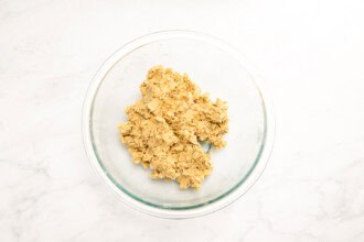 Image for recipe instruction:A glass mixing bowl containing a mound of raw cookie dough for a delicious Strawberry Rhubarb Crisp sits on a white marble surface.