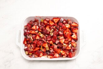 Image for recipe instruction:A white baking dish filled with chopped strawberries and rhubarb mixed with sugar, ready to be baked into a delicious Strawberry Rhubarb Crisp, sits on a white marble surface.