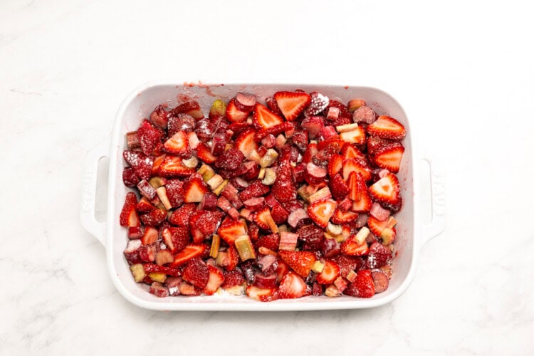 Image for recipe instruction:A white baking dish filled with chopped strawberries and rhubarb mixed with sugar, ready to be baked into a delicious Strawberry Rhubarb Crisp, sits on a white marble surface.