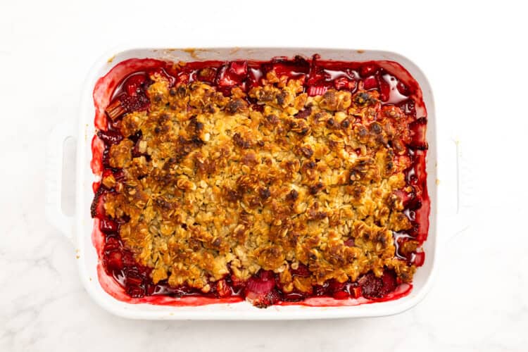A baked Strawberry Rhubarb Crisp with a golden brown oat topping in a white rectangular dish, filled with vibrant red fruit filling, photographed from above.