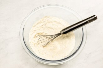 Image for recipe instruction: A glass bowl filled with flour and a metal whisk resting inside, ready for mixing the batter of an Upside Down Plum Cake, placed on a light-colored countertop.