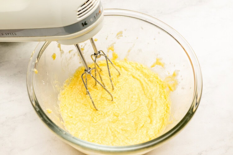 Image for recipe instruction: A hand mixer blending yellow batter for an Upside Down Plum Cake in a glass bowl on a white countertop.