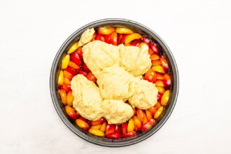 Image for recipe instruction: A round baking pan filled with sliced plums, topped with spoonfuls of raw cake dough, creates a rustic Upside Down Plum Cake, seen from above on a white background.