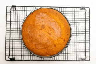 Image for recipe instruction: A round, golden-brown Upside Down Plum Cake rests on a cooling rack with a white background.