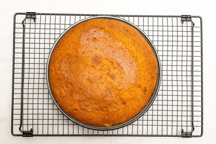 Image for recipe instruction: A round, golden-brown Upside Down Plum Cake rests on a cooling rack with a white background.