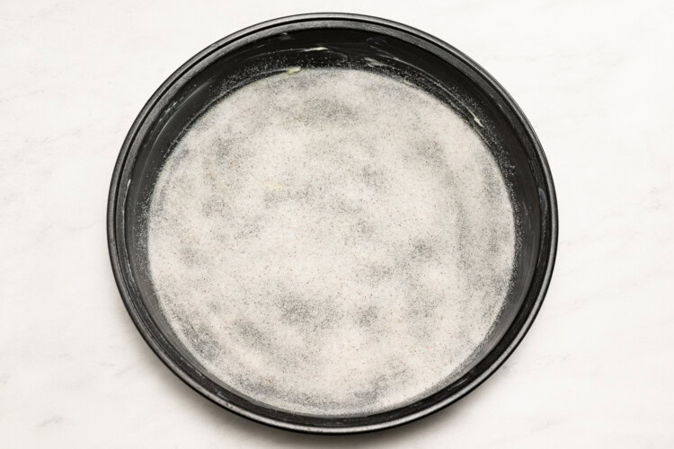 Image for recipe instruction: A round baking pan coated with a thin layer of butter and sugar, ready to create a delicious Upside Down Plum Cake, sits on a white surface.