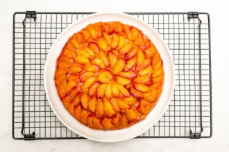 Image for recipe instruction: A round Upside Down Plum Cake with neatly arranged peach slices on top sits beautifully on a white plate, set atop a cooling rack.