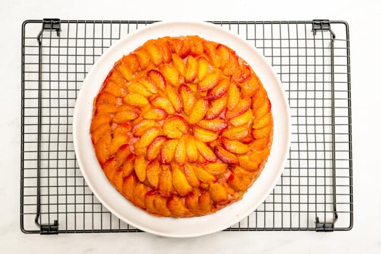 Image for recipe instruction: A round Upside Down Plum Cake with neatly arranged peach slices on top sits beautifully on a white plate, set atop a cooling rack.