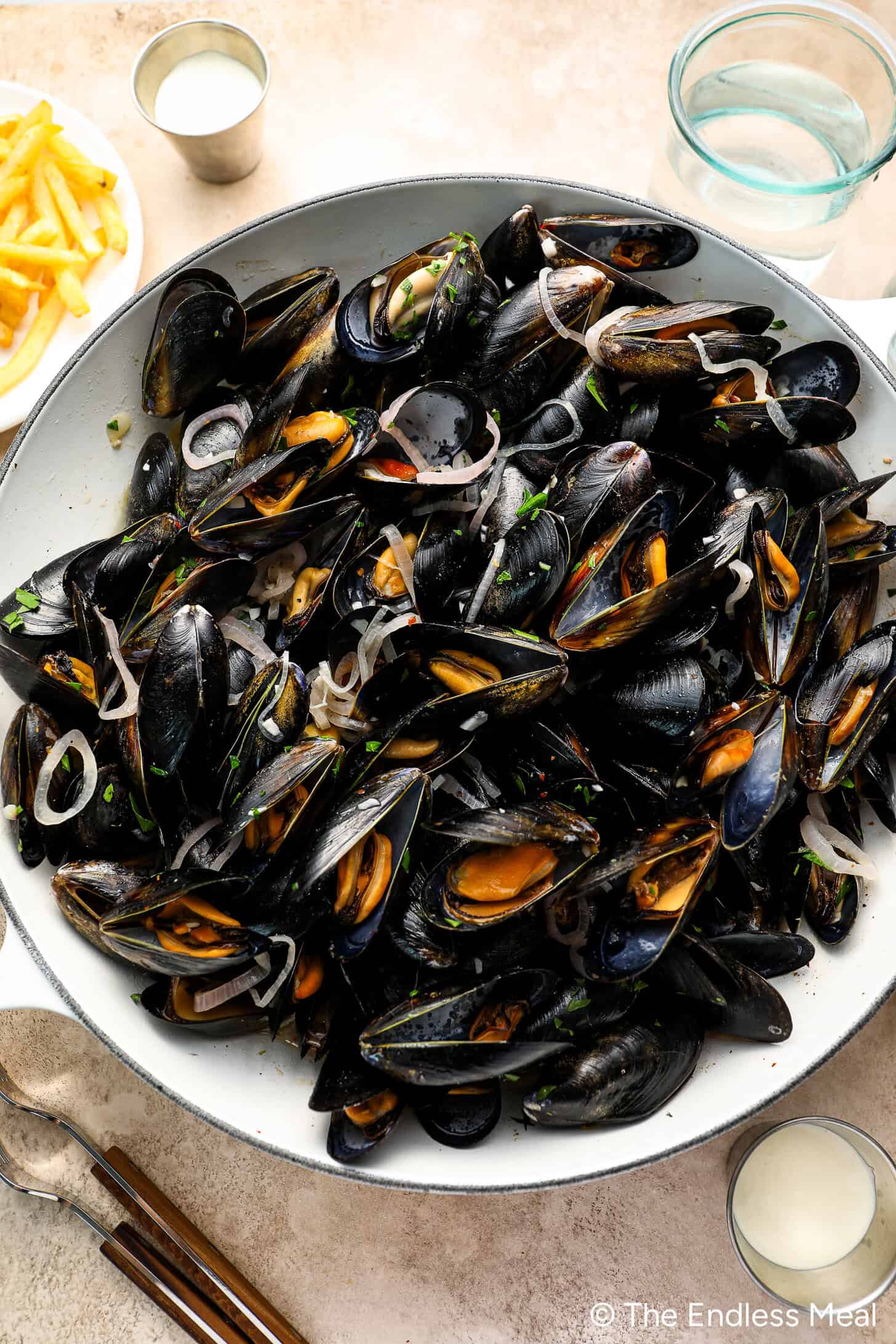 A large bowl of Moules Frites—cooked mussels garnished with sliced shallots—served alongside crispy fries, dipping sauce, and a refreshing glass of water.