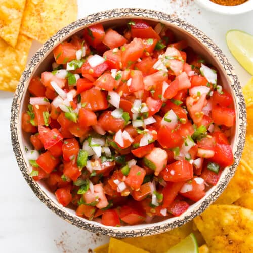 A bowl of fresh Pico de Gallo with chopped tomatoes, onions, cilantro, and jalapeños sits surrounded by crispy tortilla chips, lime wedges, and a small bowl of seasoning—perfect for snacking or topping your favorite dishes.