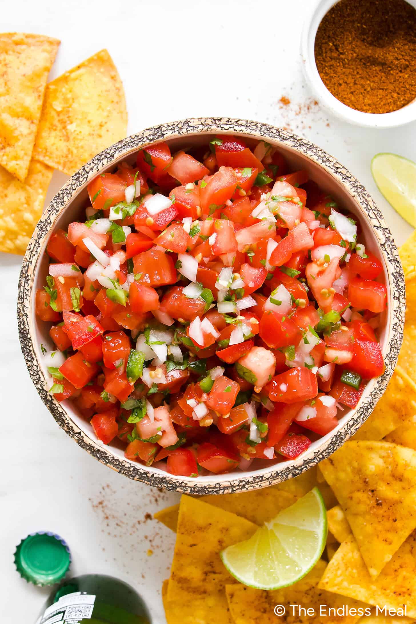 A bowl of fresh Pico de Gallo with chopped tomatoes, onions, cilantro, and jalapeños sits surrounded by crispy tortilla chips, lime wedges, and a small bowl of seasoning—perfect for snacking or topping your favorite dishes.