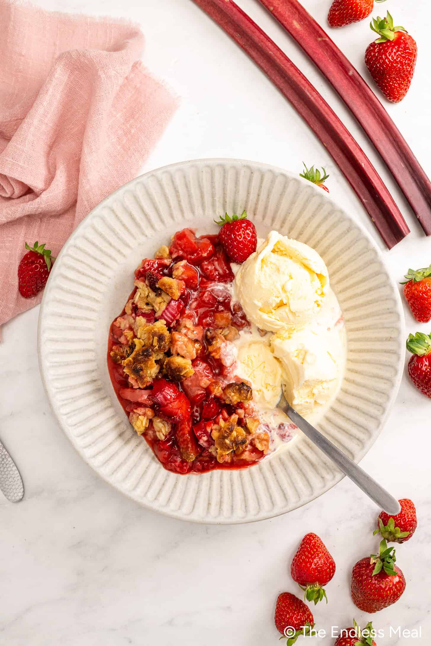 A plate with Strawberry Rhubarb Crisp and two scoops of vanilla ice cream, surrounded by fresh strawberries and rhubarb on a white surface—a perfect Strawberry Rhubarb Dessert.
