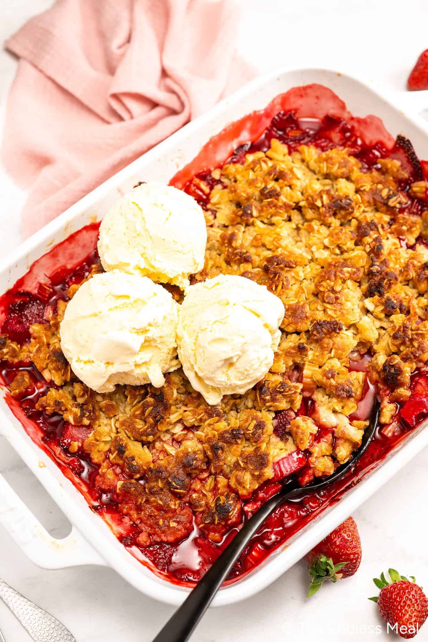 A classic Strawberry Rhubarb Crisp, this rhubarb dessert features a baked fruit filling topped with three scoops of vanilla ice cream in a white dish, served with a spoon and a pink napkin on the side.