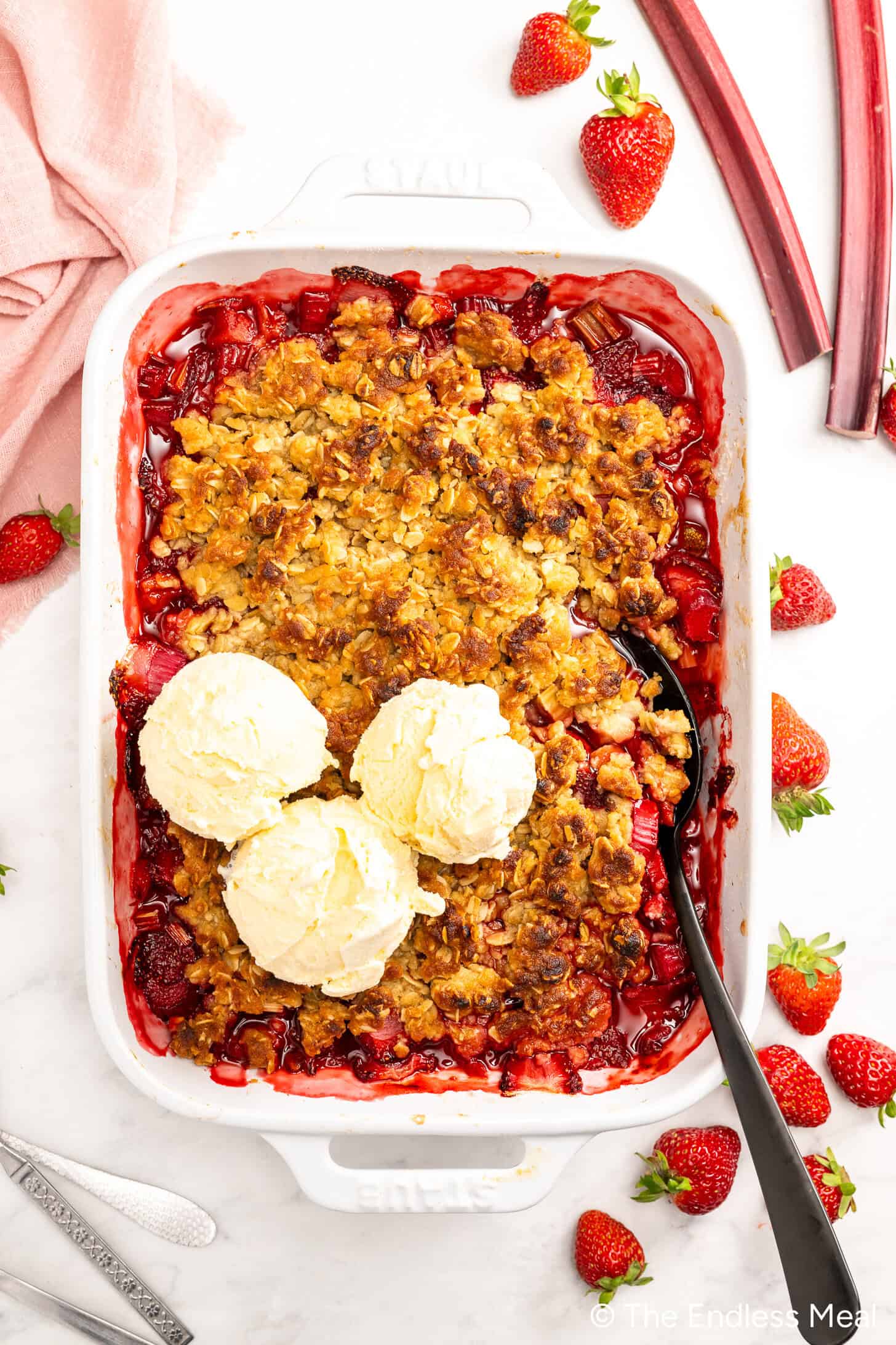 A baking dish filled with warm Strawberry Rhubarb Crisp is topped with three scoops of vanilla ice cream, accompanied by a serving spoon and fresh strawberries on the side.
