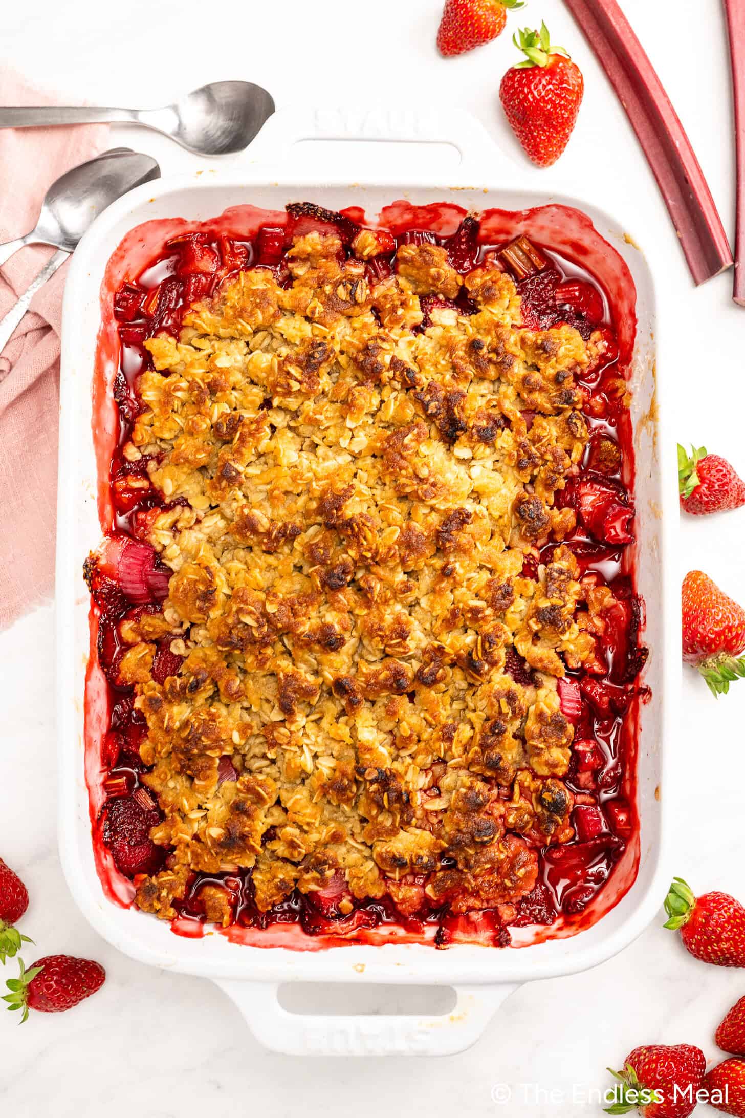 A baked Strawberry Rhubarb Crisp in a white rectangular dish, topped with a golden oat crumble, surrounded by fresh strawberries and rhubarb stalks on a white surface—a classic Strawberry Rhubarb Dessert.