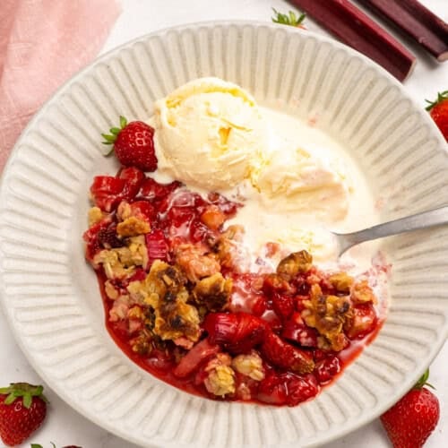 A bowl of Strawberry Rhubarb Crisp topped with vanilla ice cream, served with a spoon, fresh strawberries, and rhubarb stalks alongside.