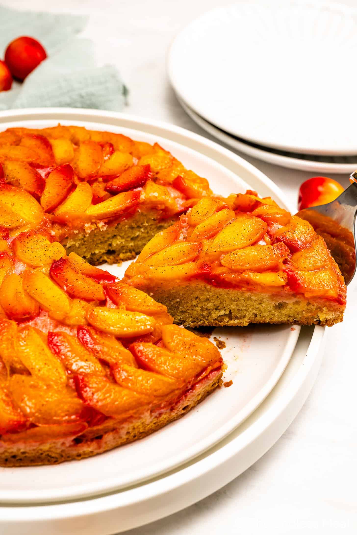 A slice of Upside Down Plum Cake is being lifted from the whole cake, which is topped with caramelized fruit slices and displayed on a white plate.
