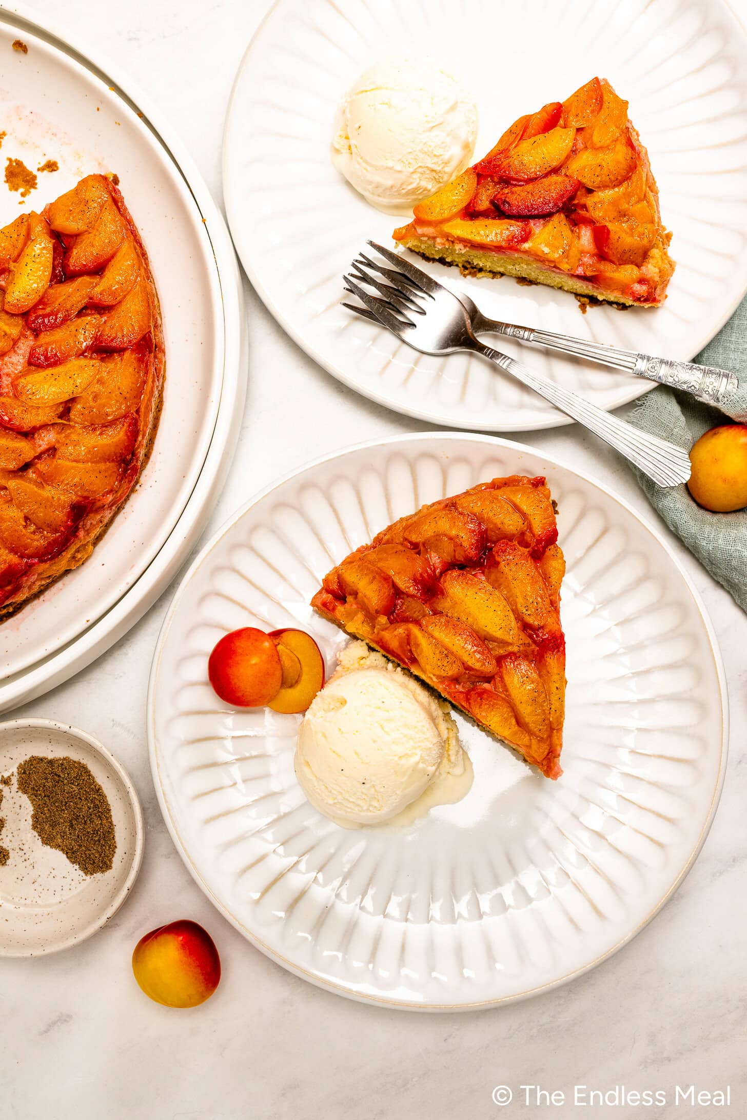 Two plates with slices of Upside Down Plum Cake served with vanilla ice cream, forks, fresh peaches, and a partial view of the cake and ground pepper on the side.