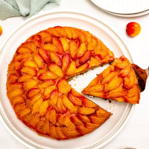 An Upside Down Plum Cake sits on a white plate, with one slice being lifted; fresh whole and halved plums are scattered around.