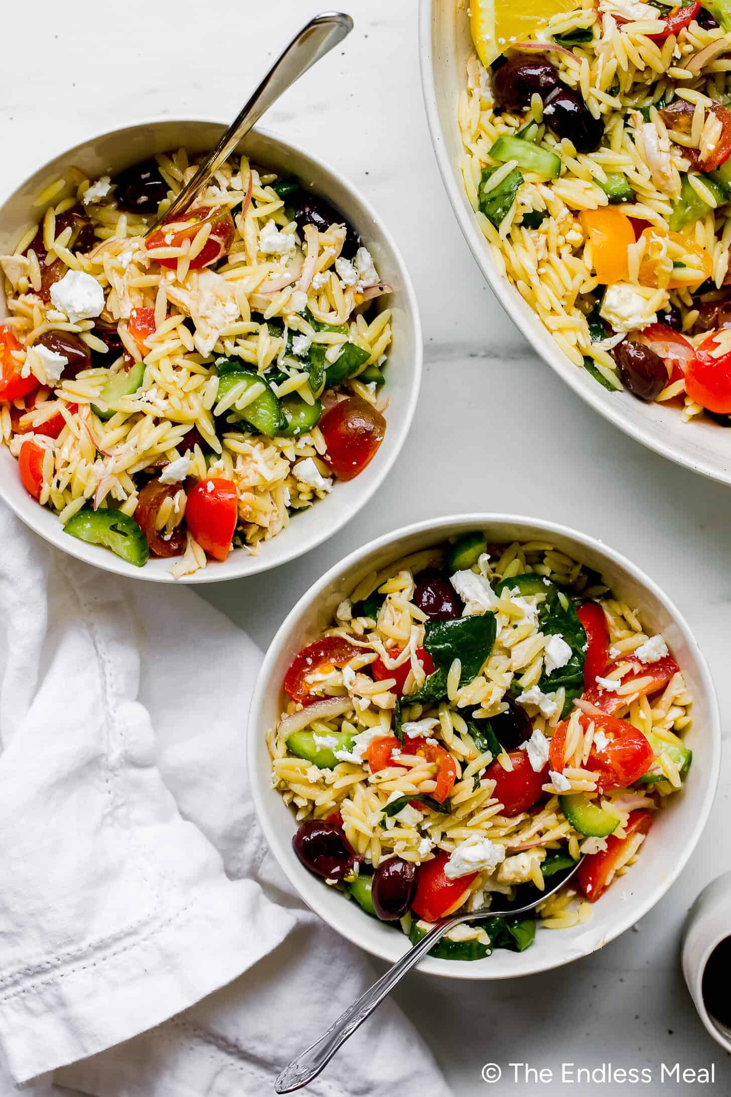Two bowls and a serving dish of Mediterranean Orzo Salad with cherry tomatoes, cucumbers, spinach, olives, and feta are placed on a white surface, each bowl topped with a spoon.