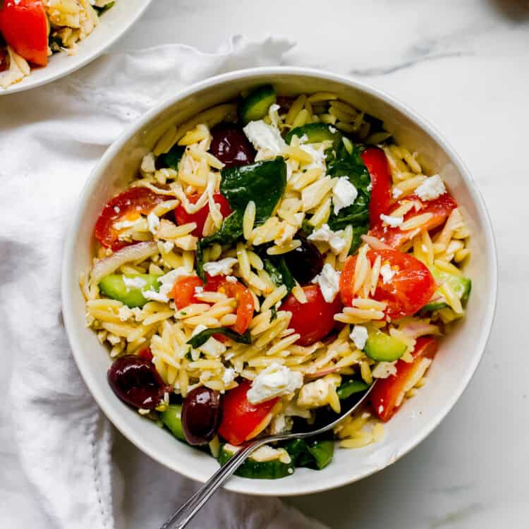 A white bowl filled with Mediterranean Orzo Salad—packed with cherry tomatoes, cucumber, spinach, olives, and feta cheese—with a fork resting inside.