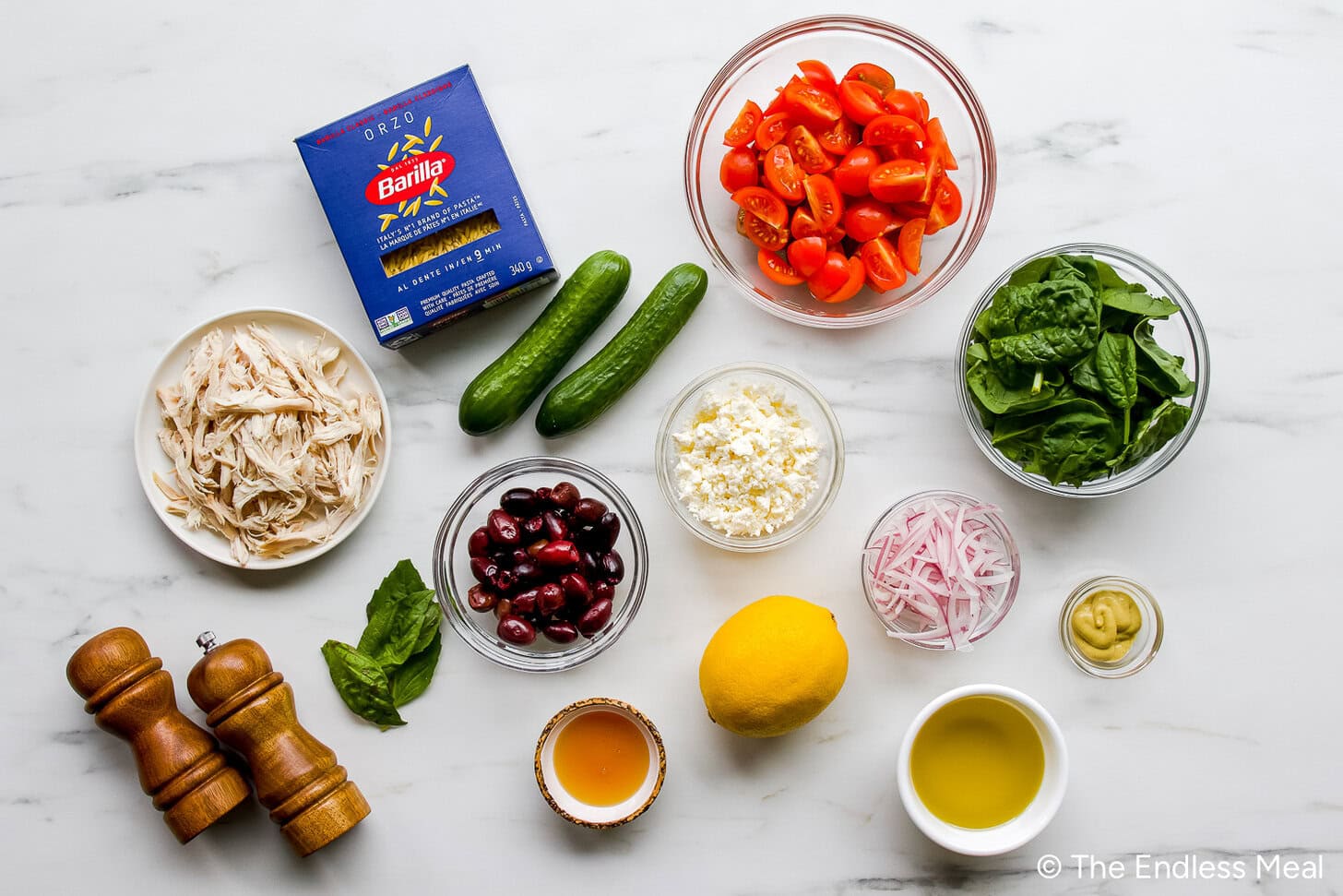 Overhead view of ingredients for a Mediterranean Orzo Salad, including orzo pasta, shredded chicken, cherry tomatoes, cucumbers, spinach, feta, kidney beans, red onion, lemon, olive oil, vinegar, and seasonings.