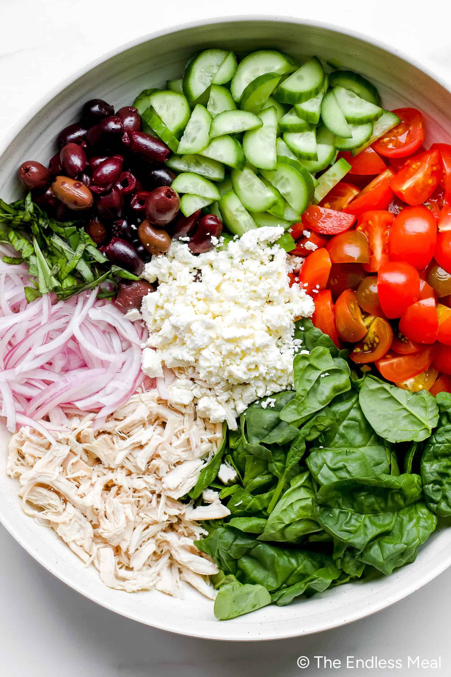 A bowl inspired by Mediterranean Orzo Salad featuring shredded chicken, spinach, sliced red onions, kalamata olives, cucumbers, cherry tomatoes, feta cheese, and fresh basil arranged in separate sections.