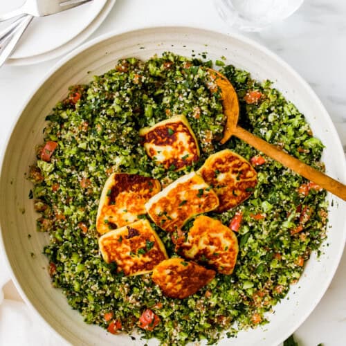 A large bowl of broccoli tabbouleh salad topped with slices of fried halloumi cheese, served with a wooden spoon. Plates, forks, and a glass of water are nearby.