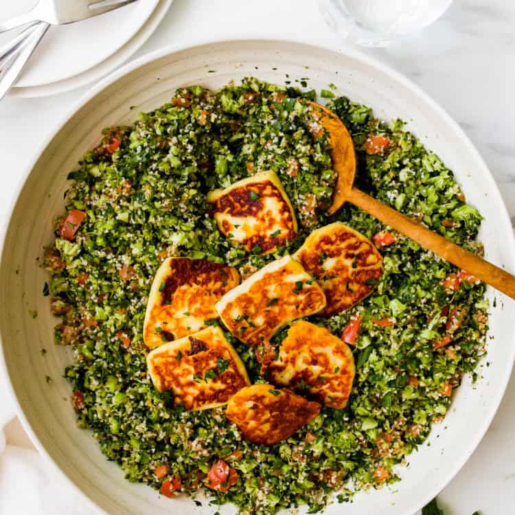 A large bowl of broccoli tabbouleh salad topped with slices of fried halloumi cheese, served with a wooden spoon. Plates, forks, and a glass of water are nearby.