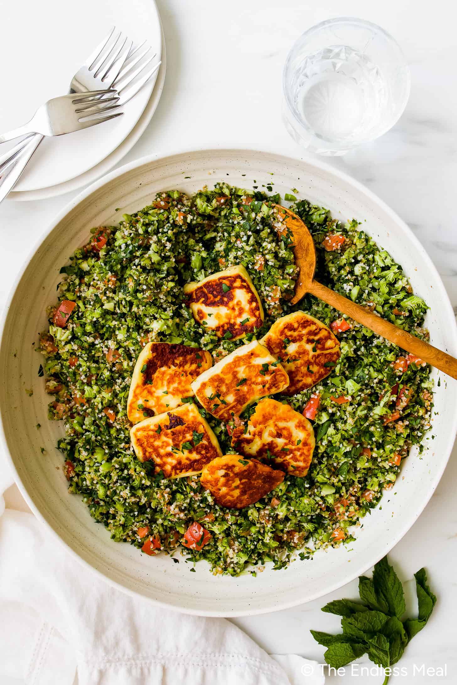 A large bowl of broccoli tabbouleh salad topped with slices of fried halloumi cheese, served with a wooden spoon. Plates, forks, and a glass of water are nearby.