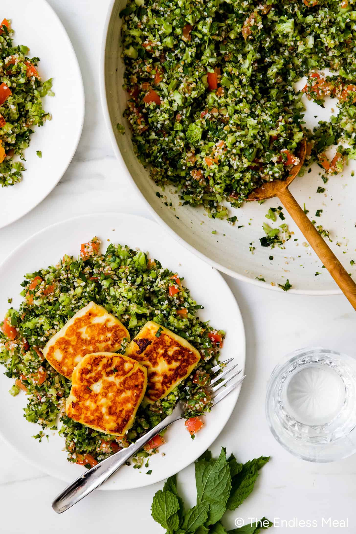 A plate of tabbouleh salad with fried halloumi slices, a fork, a glass of water, and a serving bowl of tabbouleh arranged on a white surface.