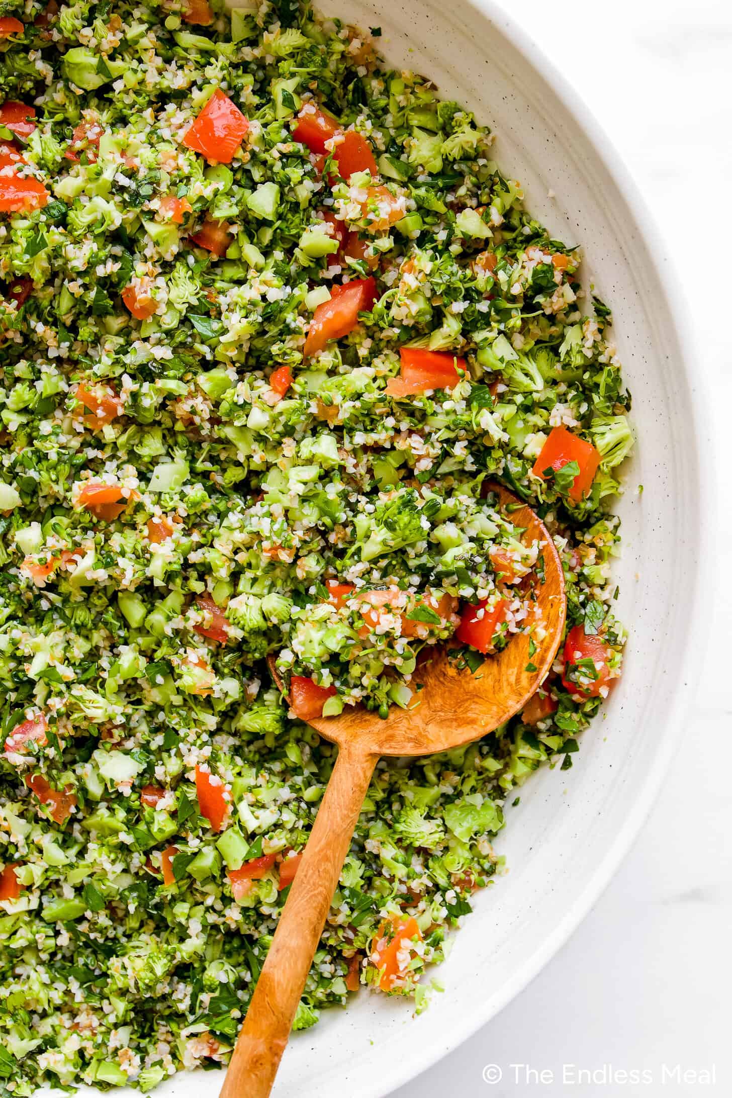 A bowl of chopped tabbouleh salad with tomatoes, parsley, and bulgur, with a wooden spoon resting inside—perfect alongside crispy fried halloumi for a fresh, flavorful meal.