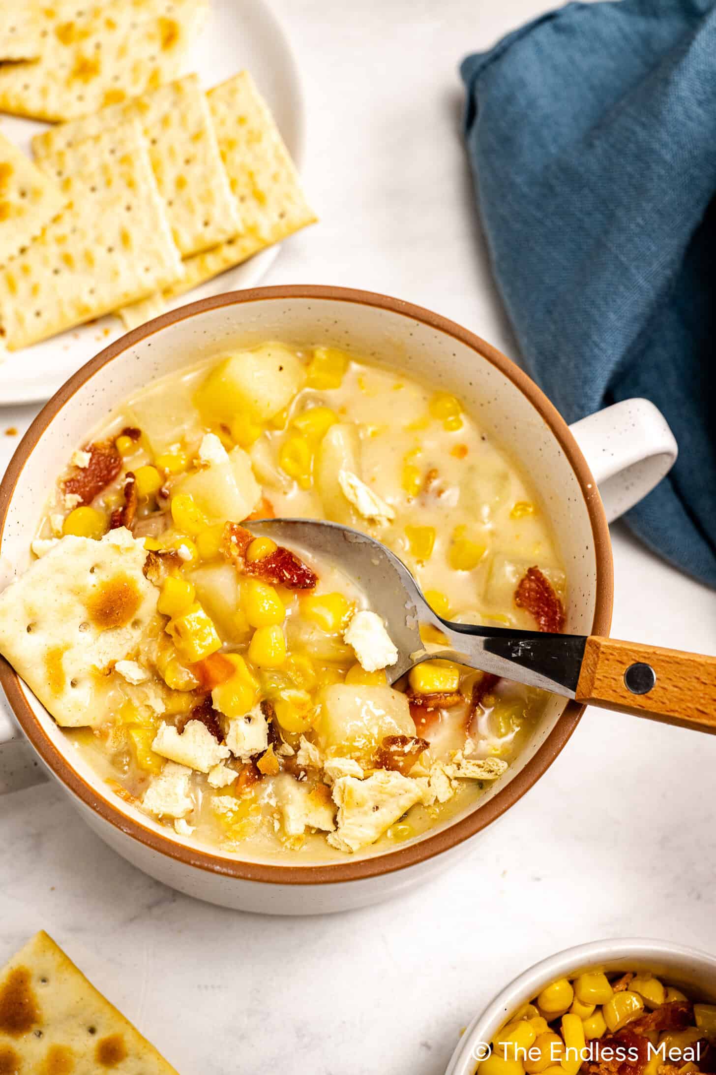 A bowl of Creamy Corn Chowder with potatoes, bacon, and crackers, with a spoon inside; extra crackers are on a plate nearby.