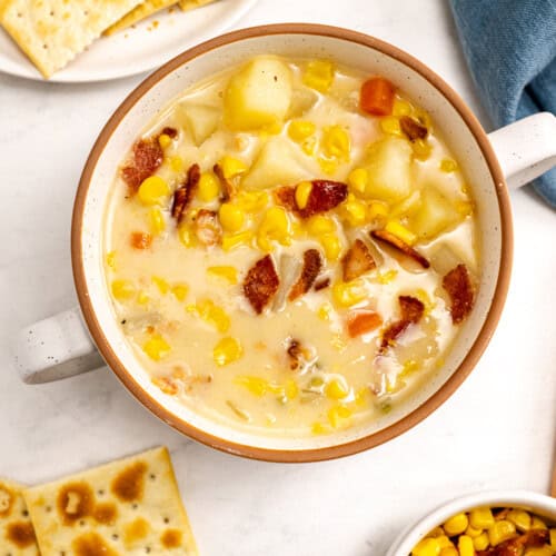 A bowl of simple corn chowder with bacon, potatoes, and carrots, served with saltine crackers on the side.