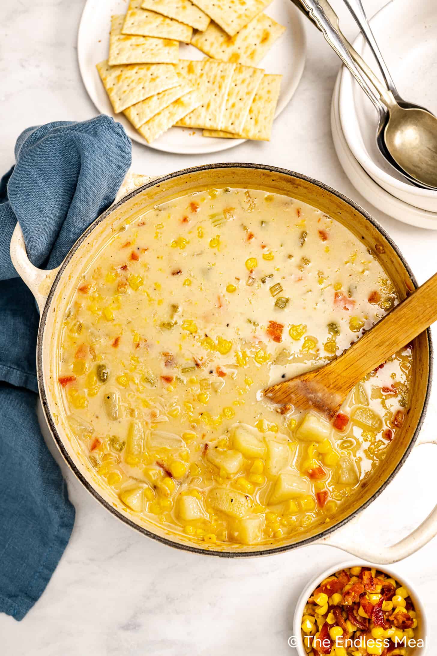 A pot of creamy corn chowder made with corn stock and fresh vegetables sits on a counter next to a plate of crackers, a blue napkin, and a bowl of toppings.