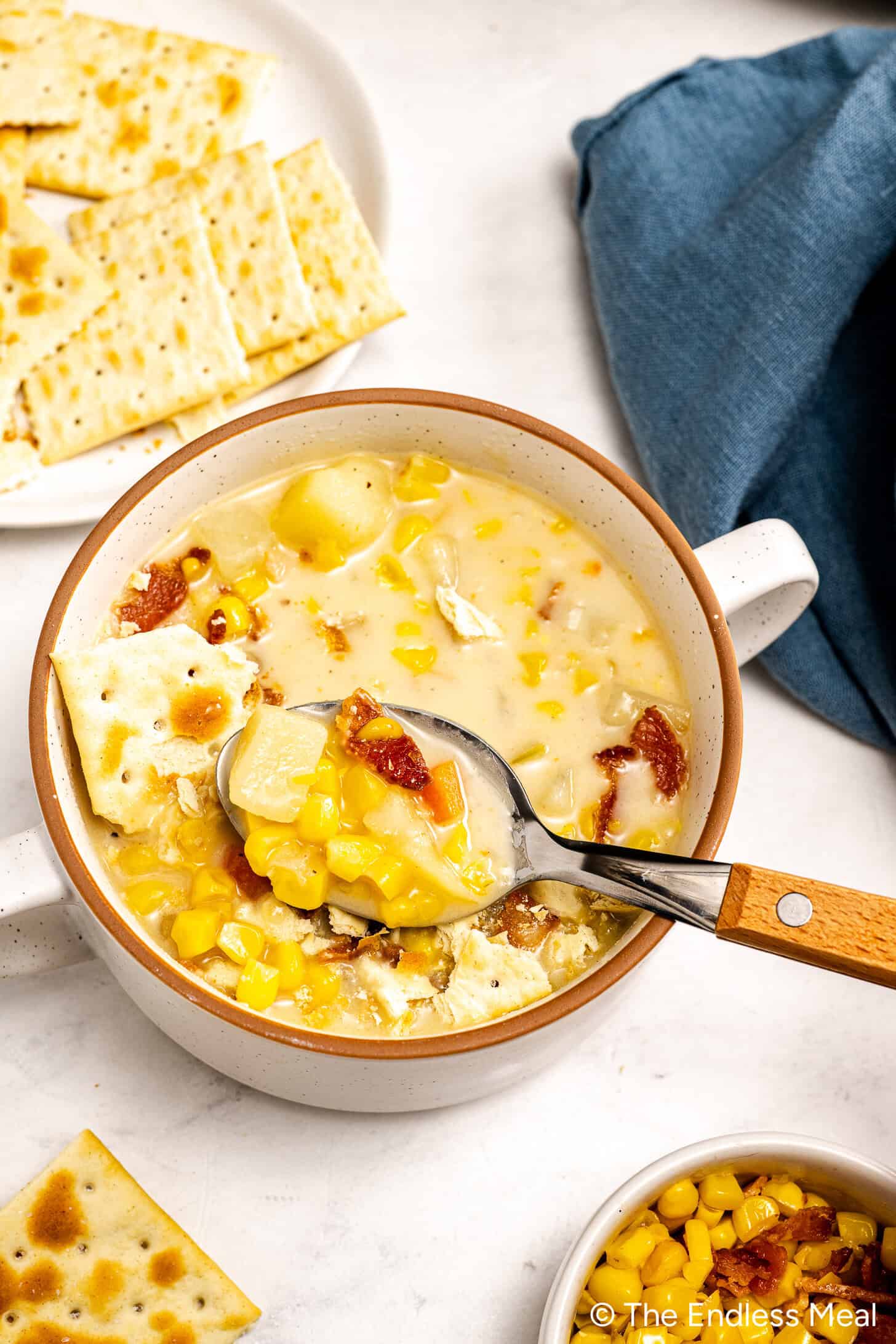 A bowl of creamy corn chowder with pieces of bacon and saltine crackers, a spoon lifting a portion; crackers and a blue napkin nearby highlight this simple corn chowder made with rich corn stock.