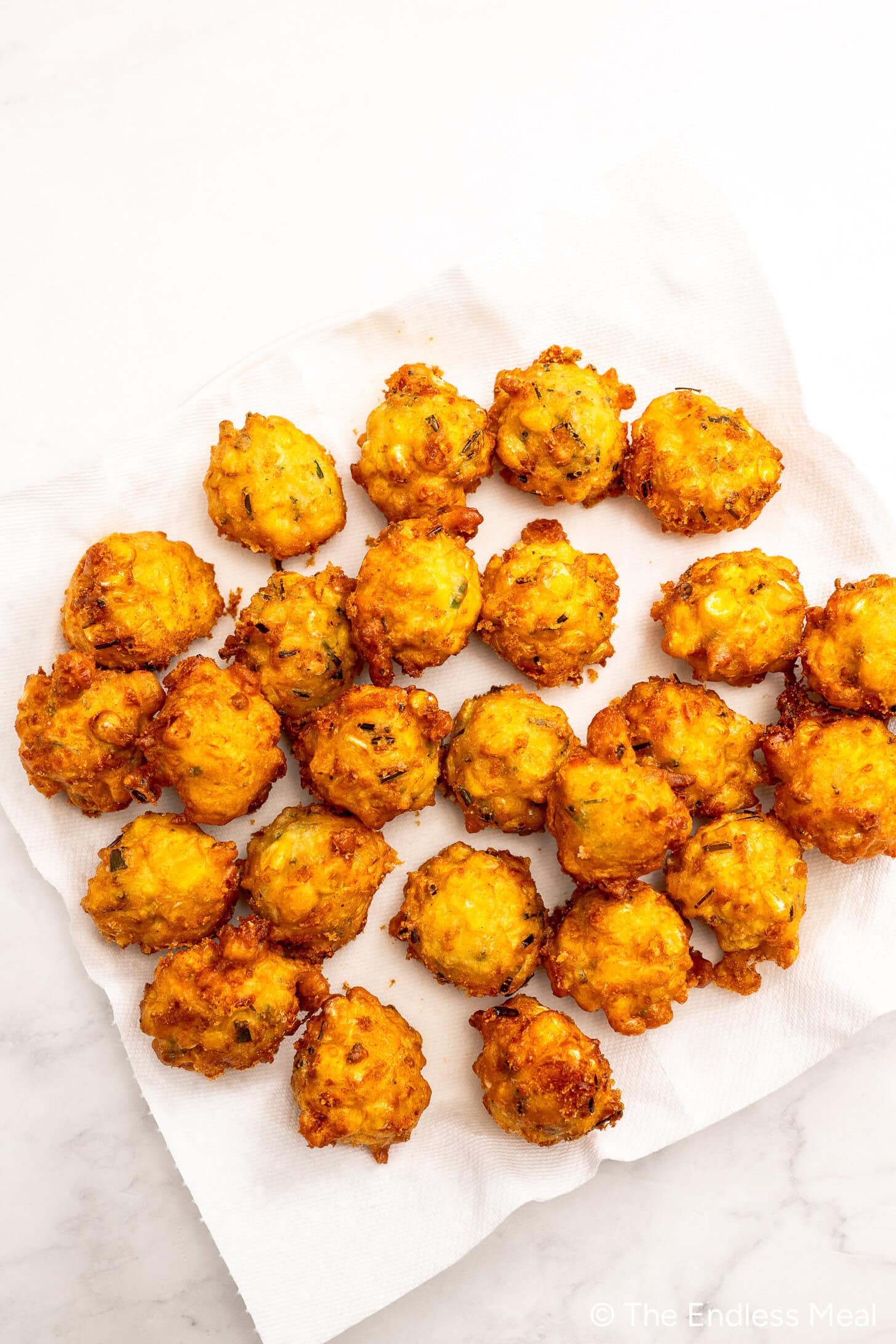 Twenty golden-brown corn fritters rest on a white paper towel on a light surface, arranged in rows and closely grouped together—the perfect addition to your favorite corn recipes.