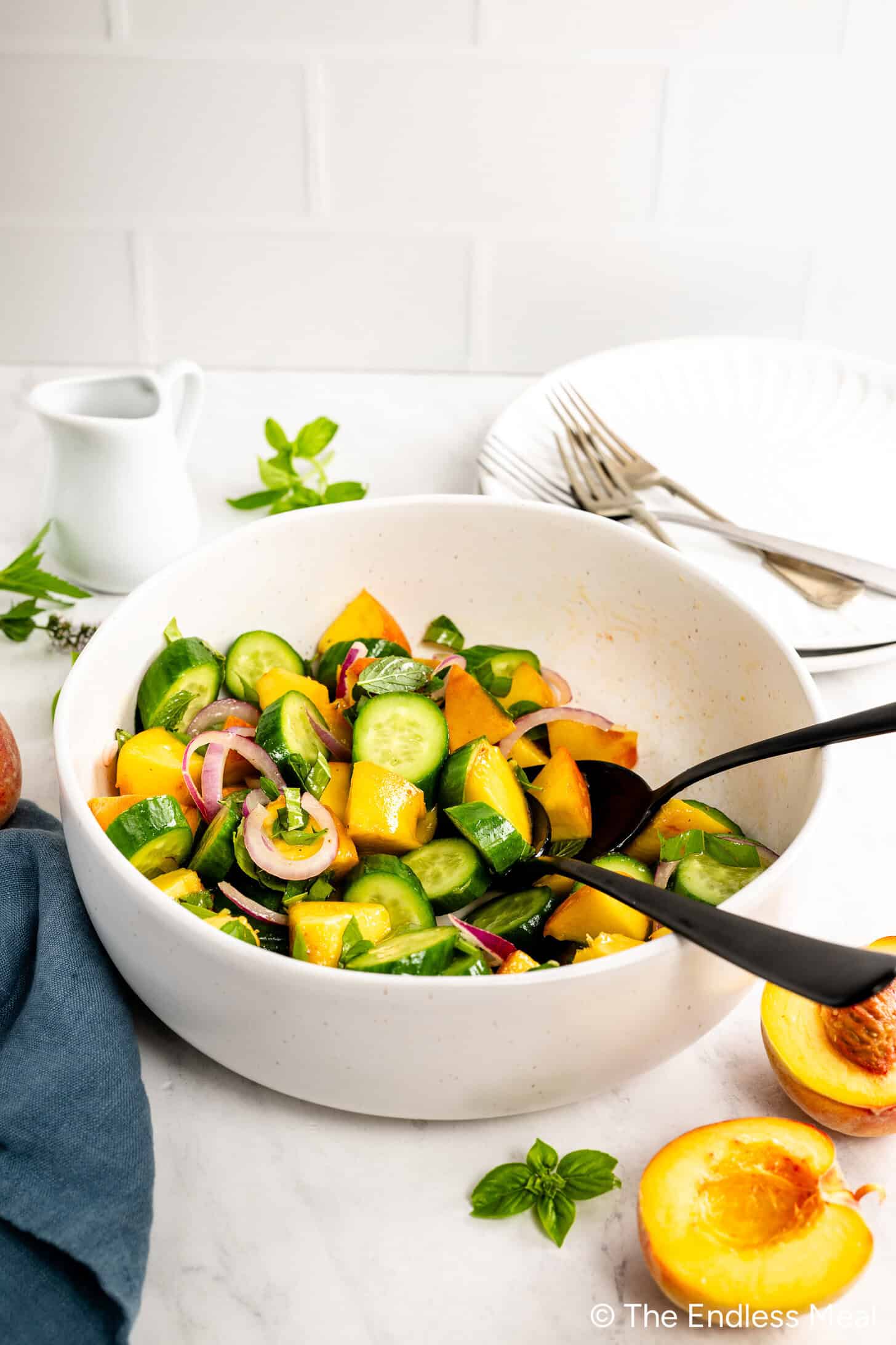 A white bowl filled with Cucumber Peach Salad—cucumbers, peaches, red onions, and herbs—sits on a marble counter with serving utensils, plates, and a small pitcher of Hot Honey Vinaigrette nearby.