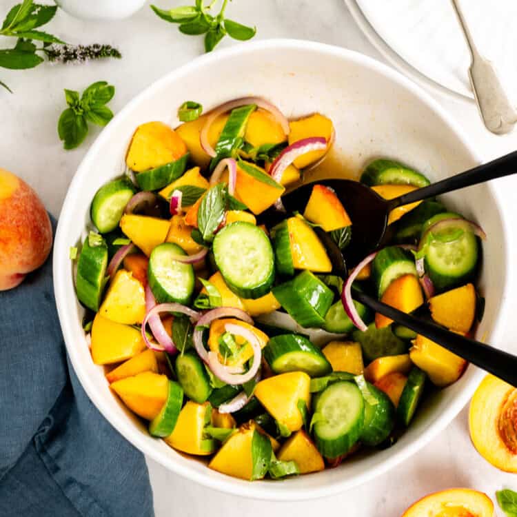 A bowl of Cucumber Peach Salad with sliced cucumbers, peaches, red onions, and fresh herbs is served alongside a small pitcher of Hot Honey Vinaigrette, with tongs and plates with forks nearby.