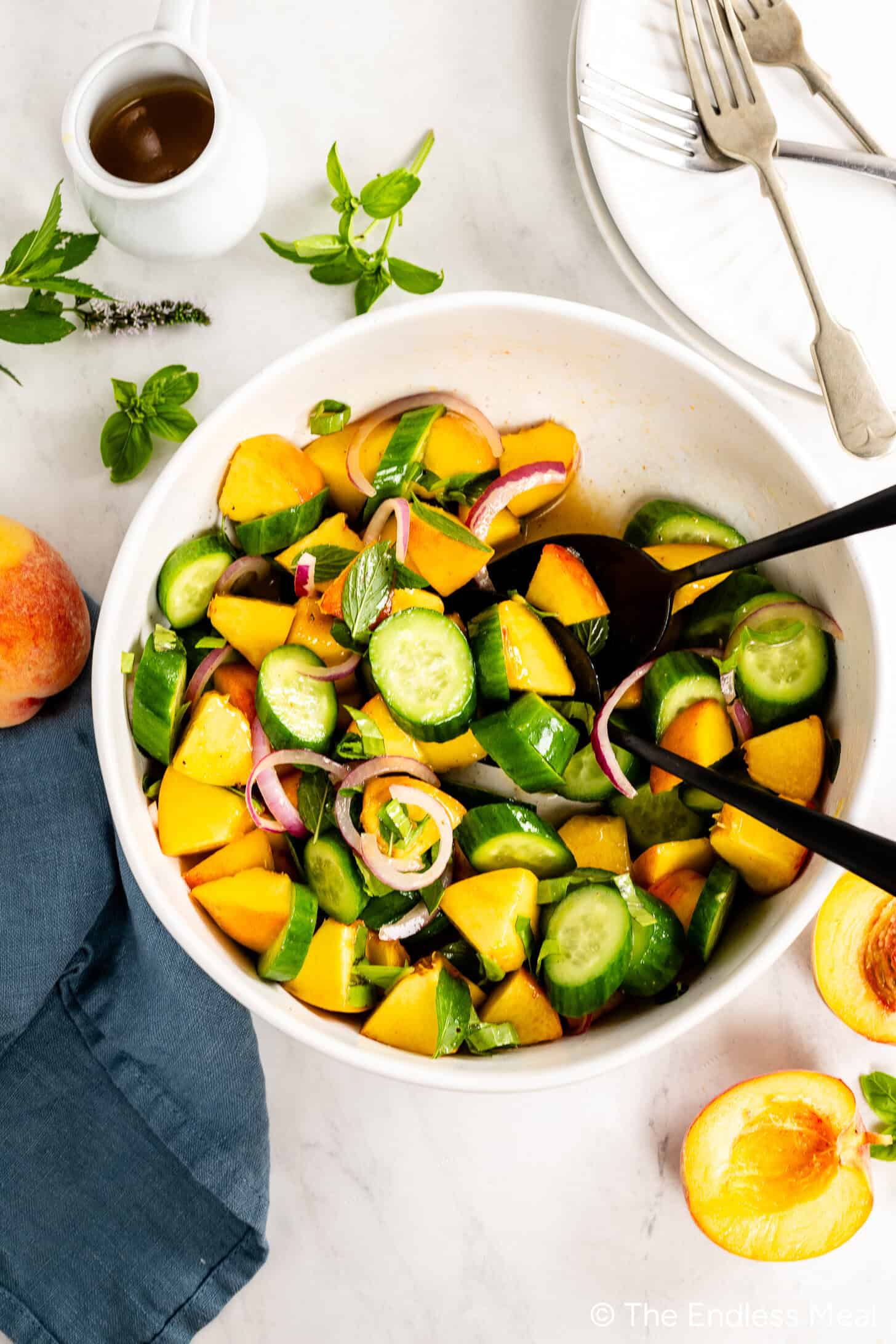 A bowl of Cucumber Peach Salad with sliced cucumbers, peaches, red onions, and fresh herbs is served alongside a small pitcher of Hot Honey Vinaigrette, with tongs and plates with forks nearby.
