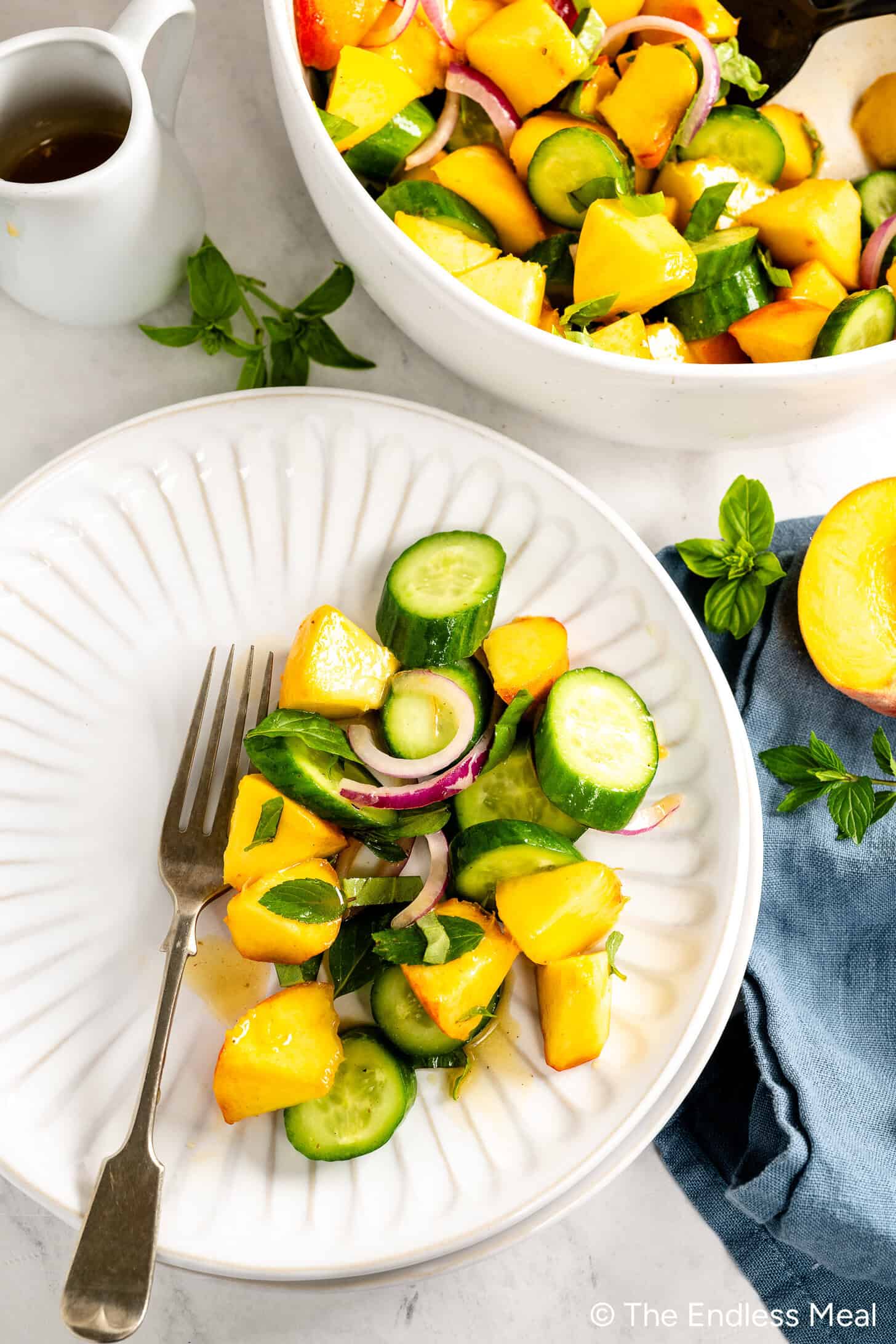 A plate with a fork holds a refreshing Cucumber Peach Salad of sliced cucumbers, peaches, and red onions. Nearby, a larger bowl with the same salad and a small pitcher of Hot Honey Vinaigrette sit on the table.